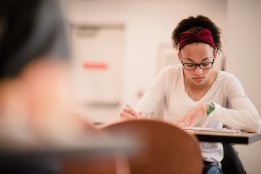 A young women wearing glasses and a white shirt sitting at a student desk working on an assignment.